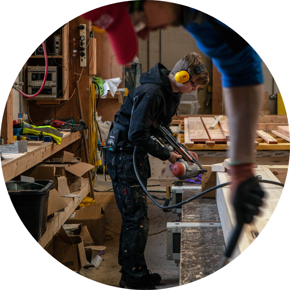 Man using power tool, uearing ear defenders in a workshop, at a work bench surrounded by wood and tools