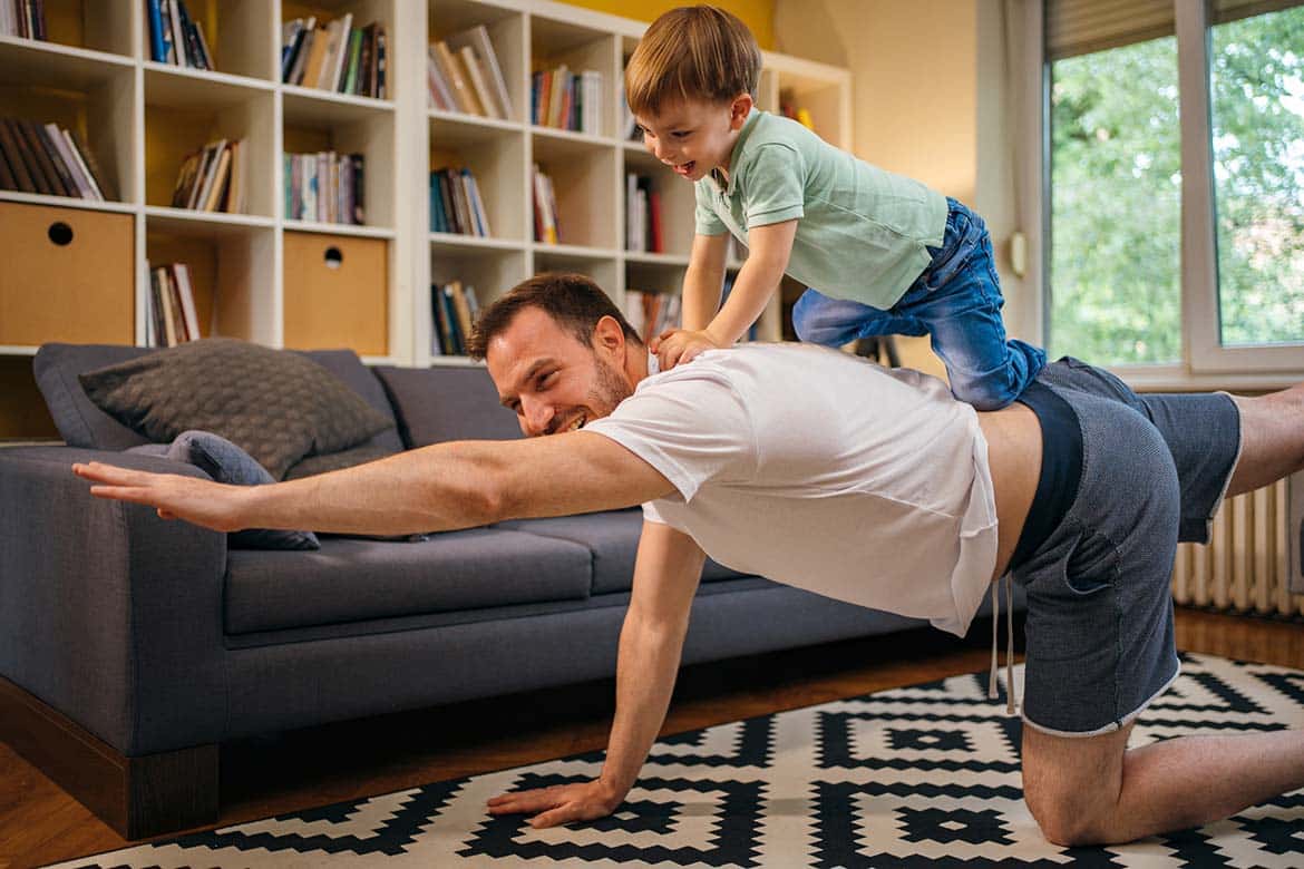 Man in white t-shirt, grey shorts on the floor in a yoga position with small boy child on his back
