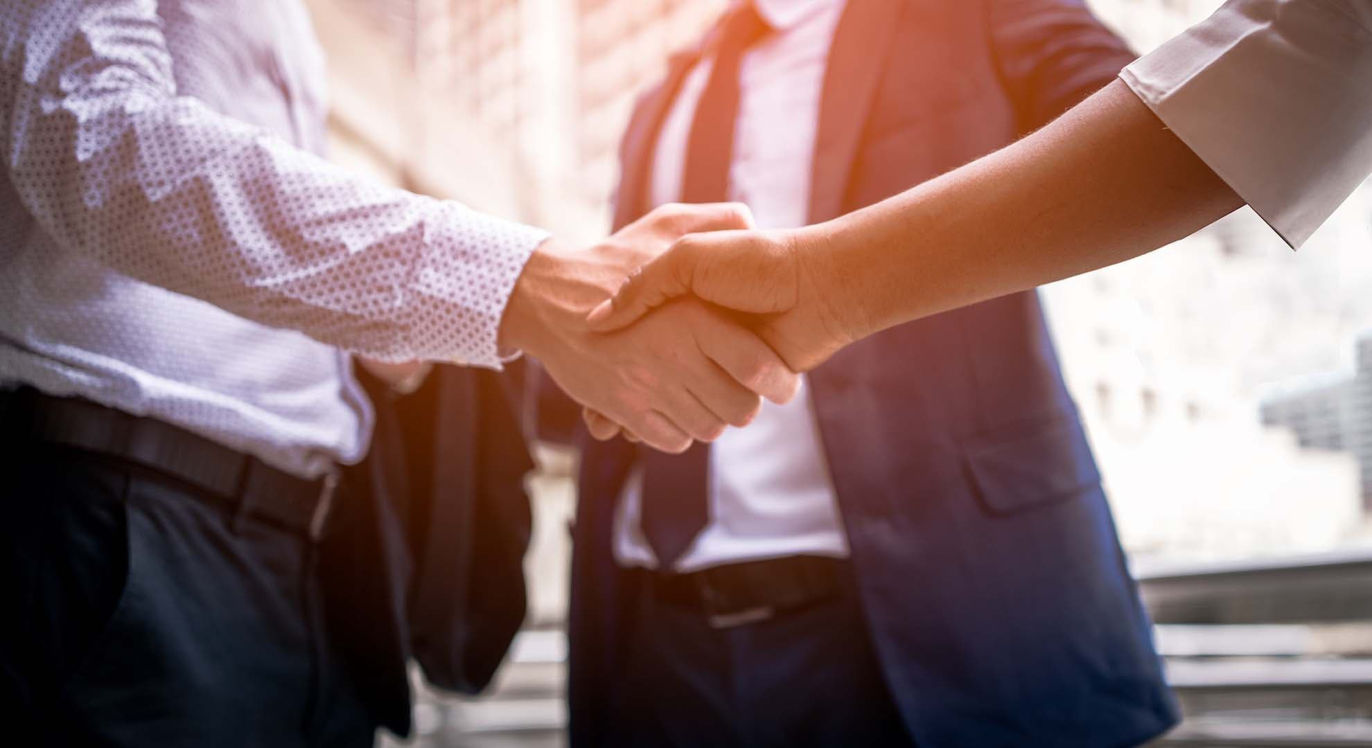 Two people shaking hands in front of a third person, in a business meeting