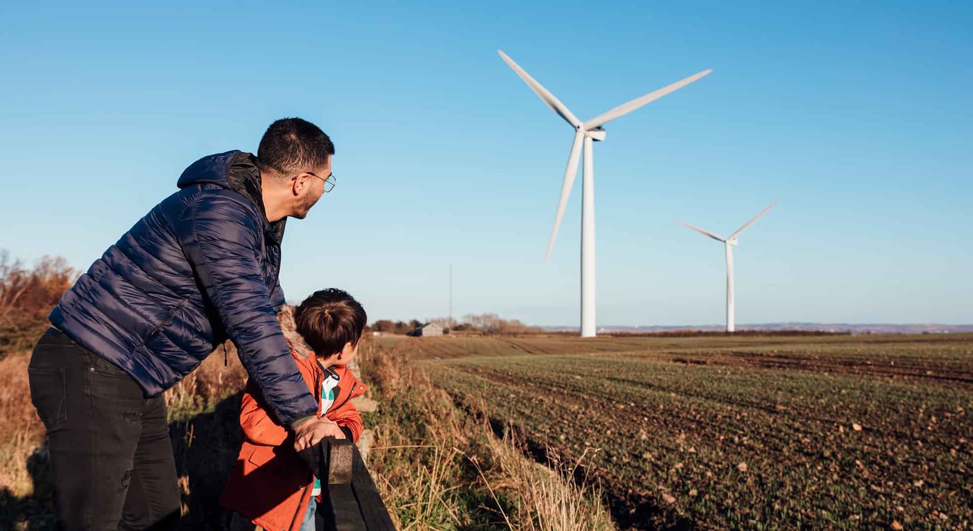 A father and son leaning against a fence in a field together, they are looking at the wind turbines.